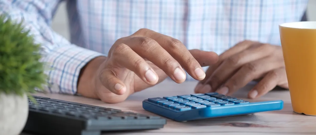 A man using a calculator at his desk next to a keyboard and coffee mug