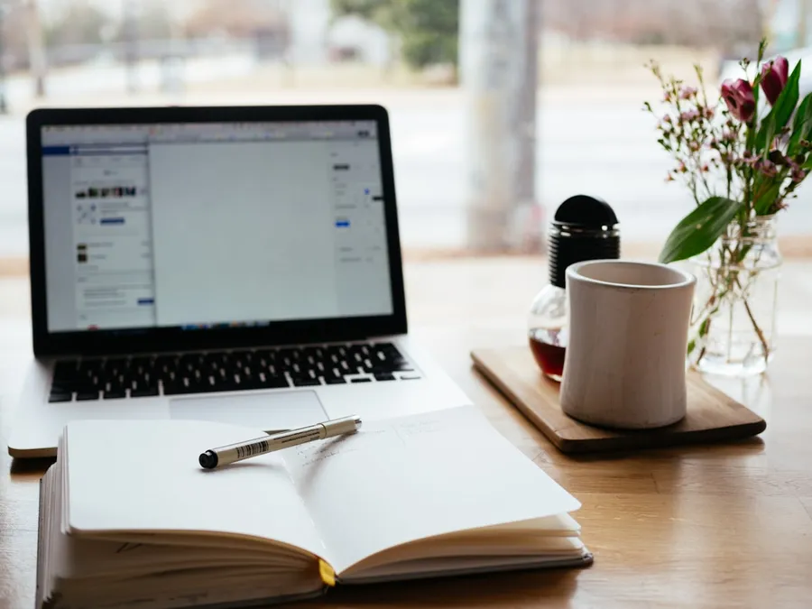 An open notebook and laptop on a desk with coffee and flowers