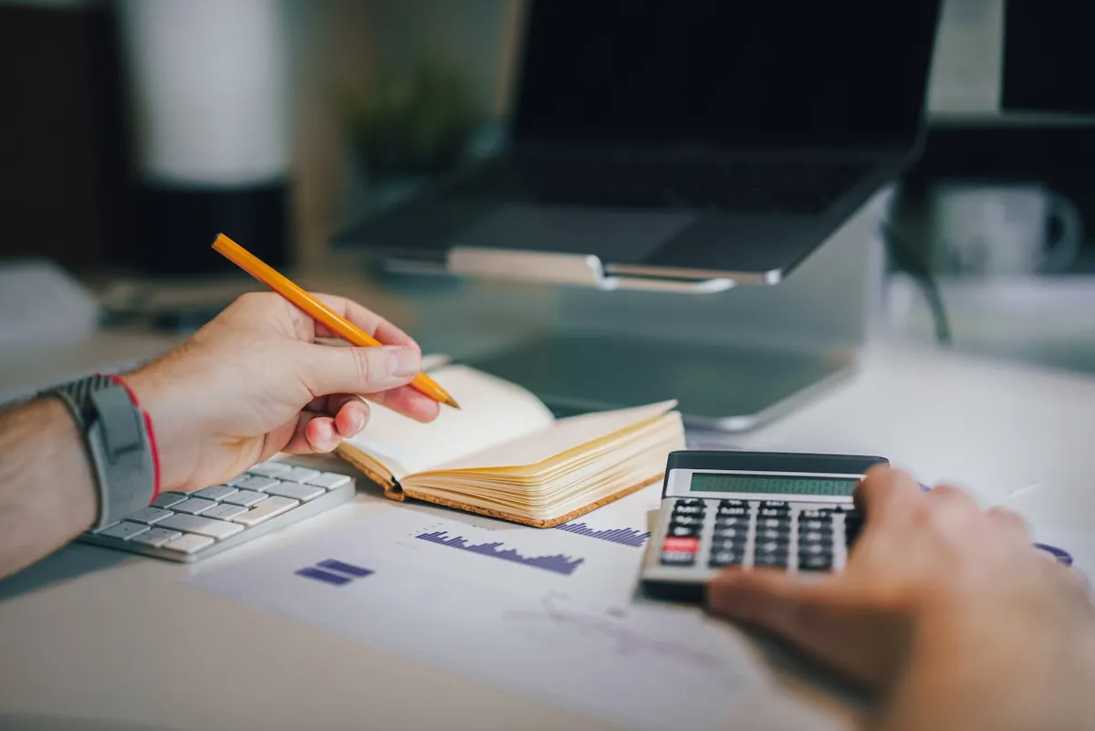 Hands using a calculator next to a notebook and laptop with a financial chart